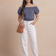 Woman wearing a blue off-shoulder top and white pants, holding a brown bag against a beige background