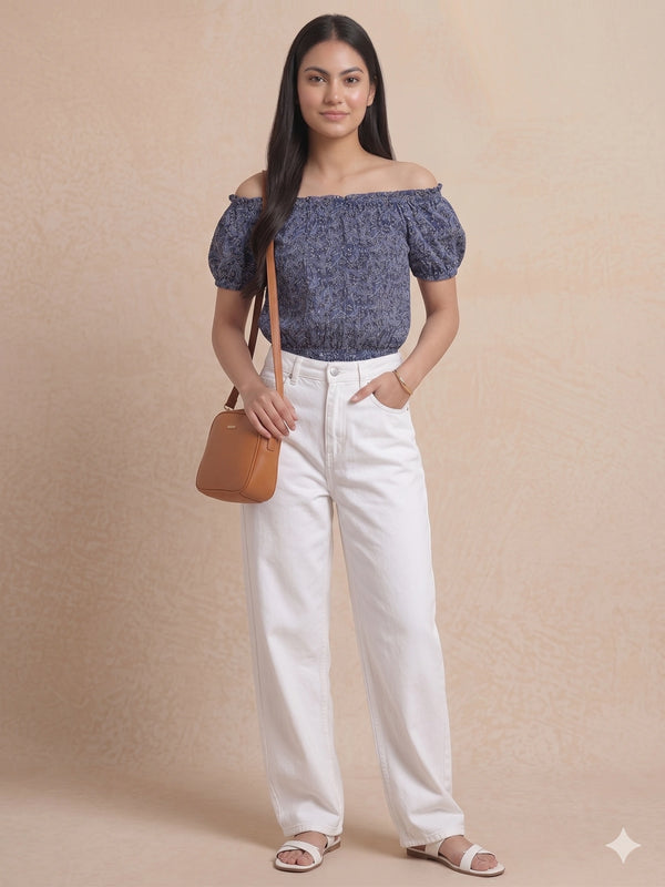 Woman wearing a blue off-shoulder top and white pants, holding a brown bag against a beige background