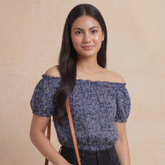 Woman wearing a blue floral off-shoulder top and holding a brown bag against a beige background