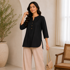 Woman in black top and beige pants standing in a room with a plant and chair.