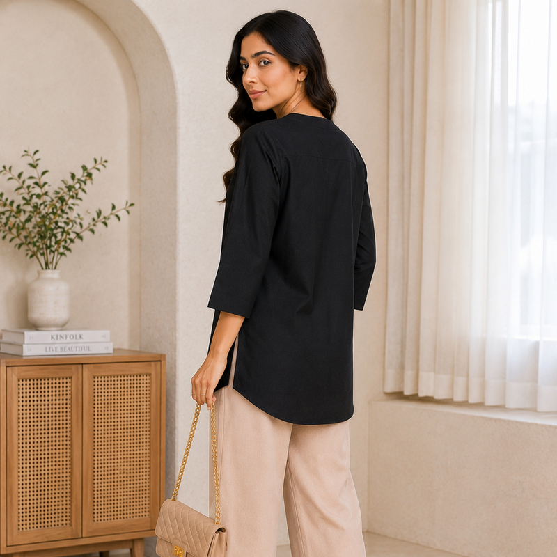 Woman in black top and beige pants holding a beige handbag in a room with a wooden cabinet and plant.