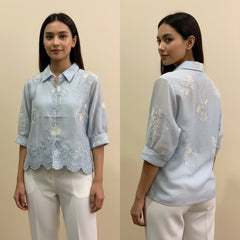 Woman wearing a light blue embroidered blouse with white pants on a beige background