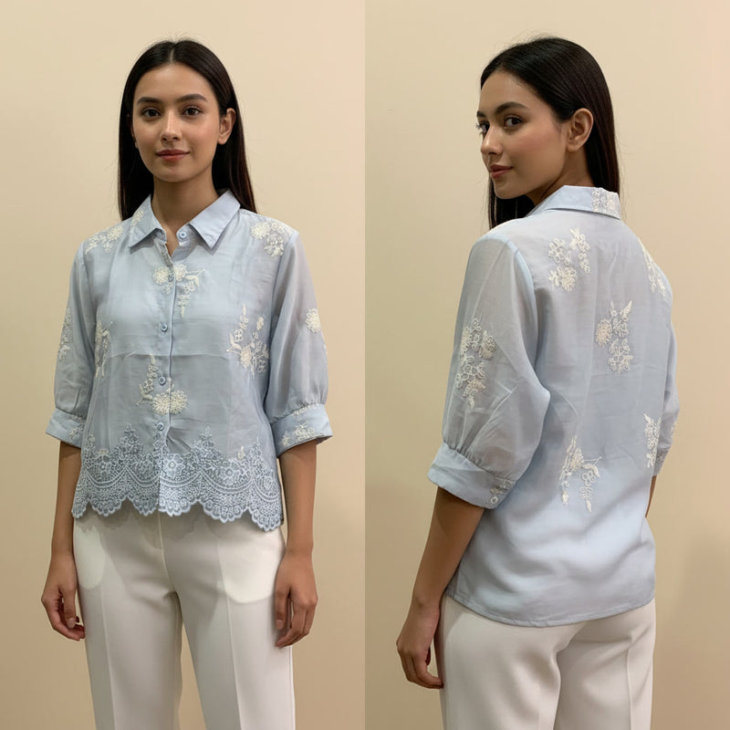 Woman wearing a light blue embroidered blouse with white pants on a beige background