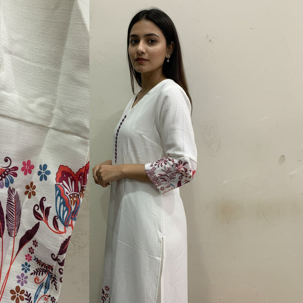 Woman in a white outfit with floral patterns standing next to fabric with similar designs.